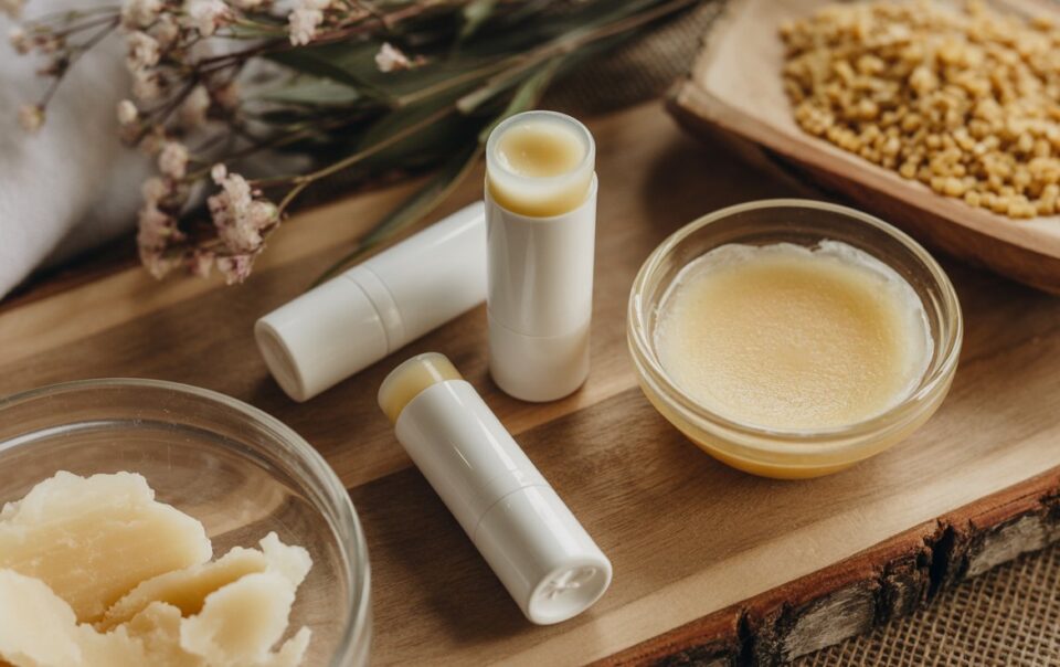 Homemade tallow lip balms in white tubes, a small glass bowl of balm, beeswax pellets, and raw shea butter displayed on a wooden board with dried flowers in the background.
