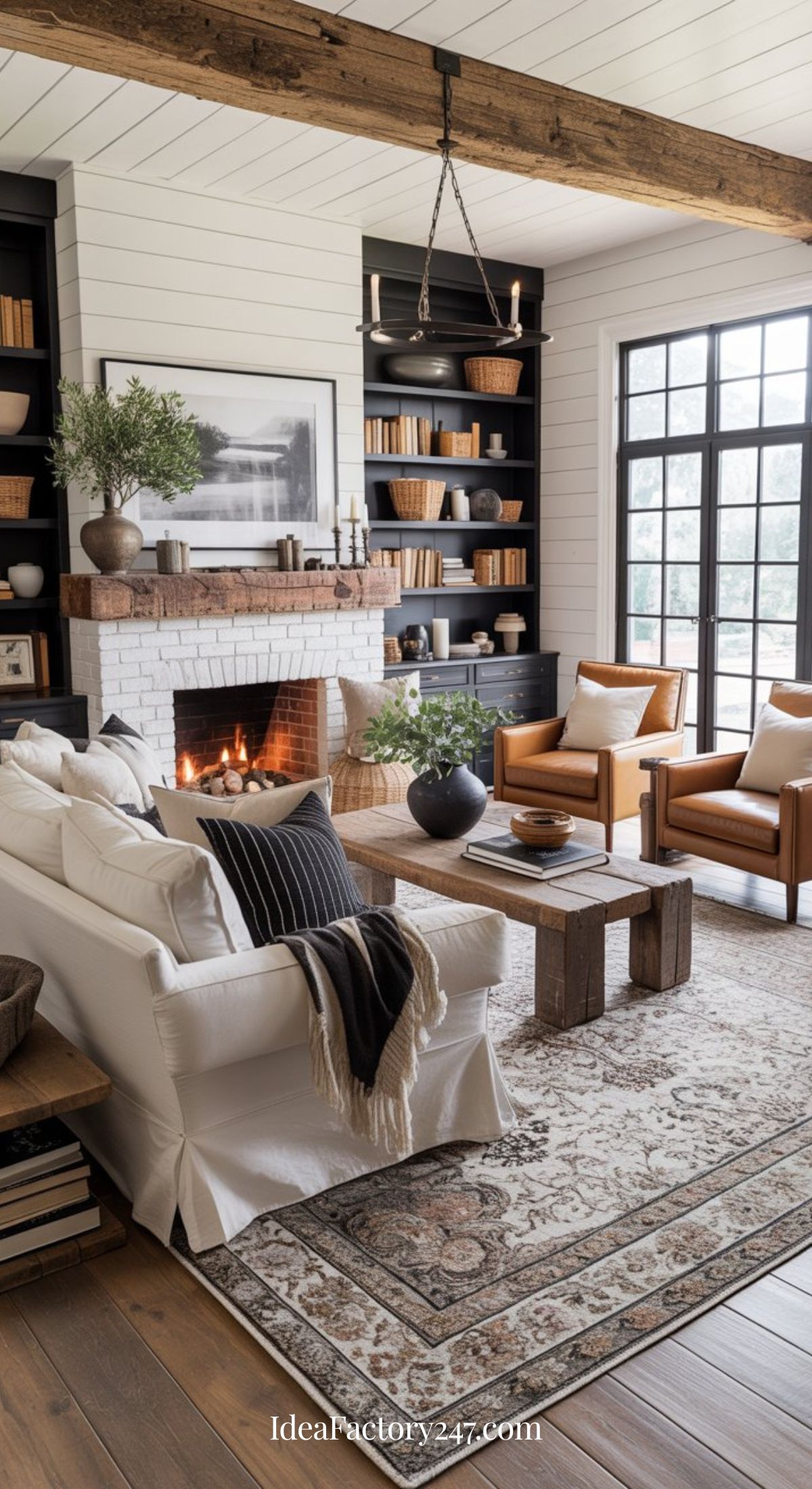 A cozy living room with a white sofa, brown armchairs, a lit fireplace with a white brick surround, built-in shelves with books and decor, large windows, and a patterned rug on a wooden floor.