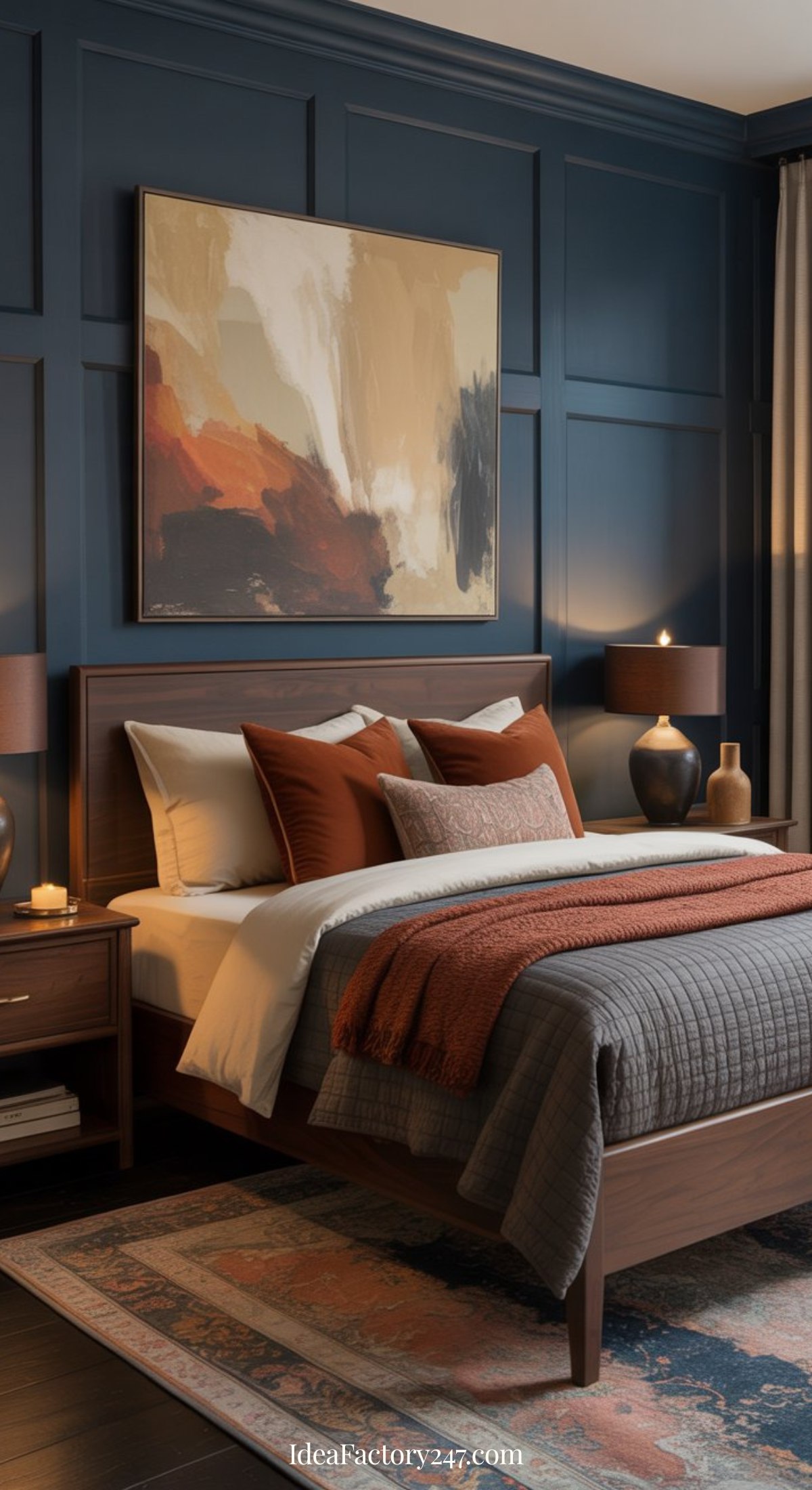 A modern blue and terracotta bedroom with dark blue paneled walls, a large abstract painting above a wooden bed, rust and brown accent pillows, gray and rust blankets, two bedside tables with lamps, and a patterned rug.