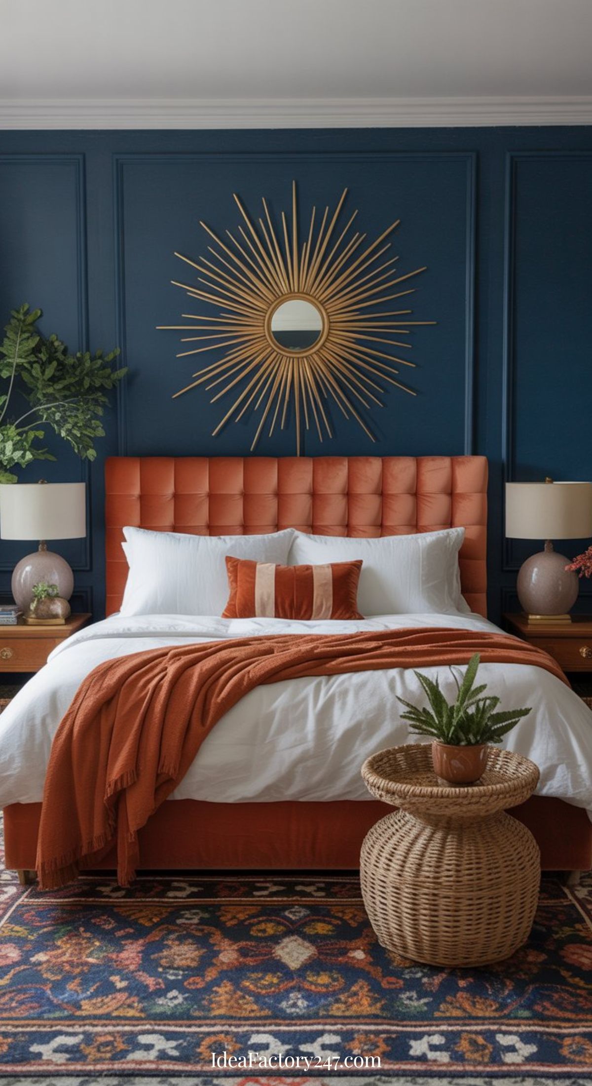 A stylish blue and terracotta bedroom with a dark blue paneled accent wall, an orange tufted headboard, white bedding with orange pillows and throw, sunburst mirror, wooden nightstands, table lamps, and a woven basket with a plant.