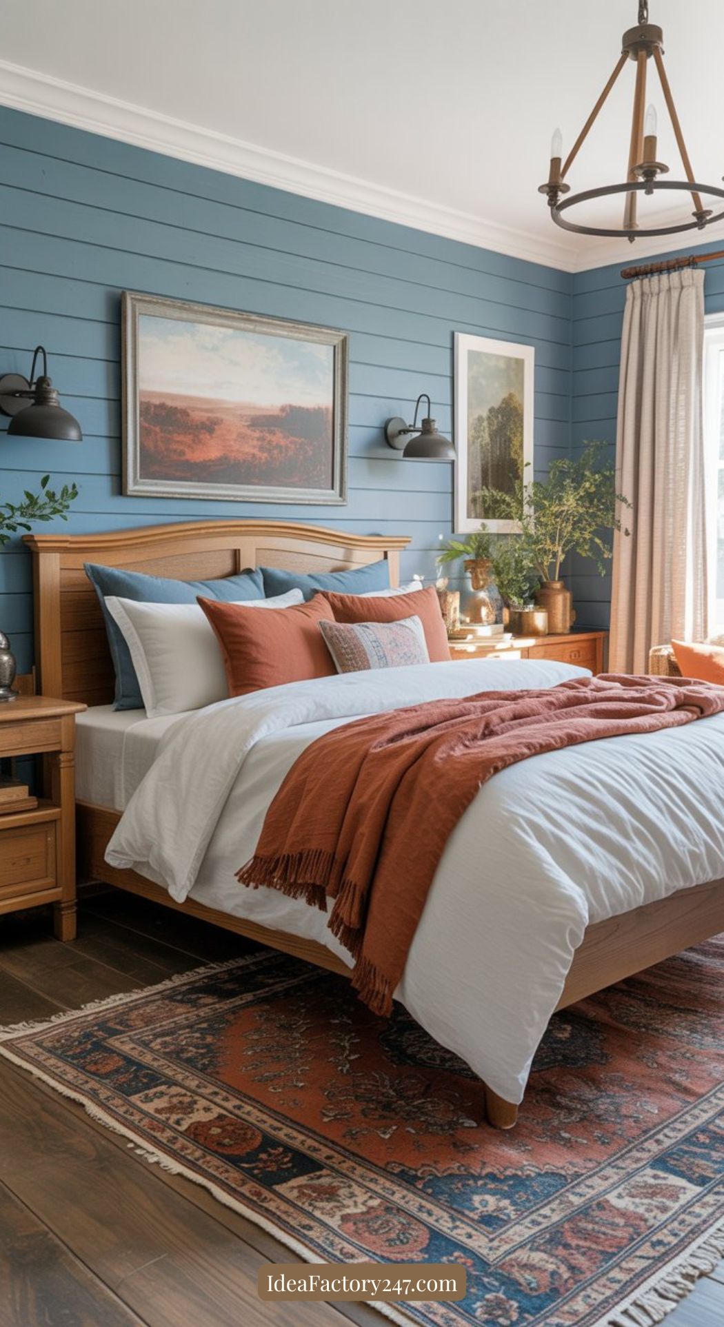 A cozy bedroom with a wooden bed, white bedding, and orange pillows and blanket. Blue shiplap walls, framed art, wall lamps, houseplants, and wooden nightstands complete the space. A patterned rug covers the floor.