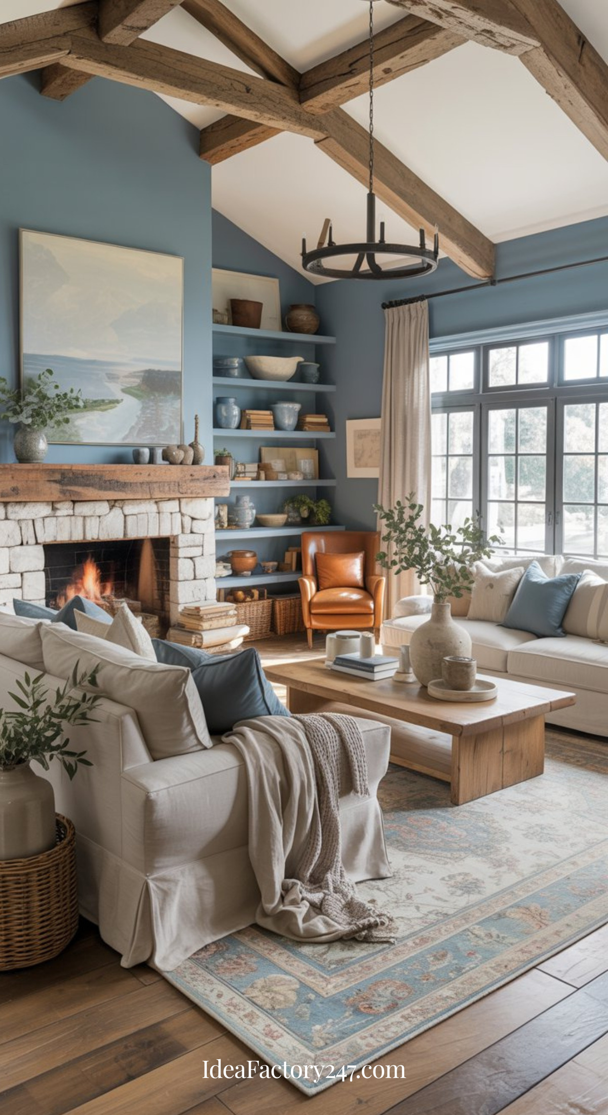 A cozy farmhouse living room with a light stone fireplace, blue walls, wooden ceiling beams, neutral sofas, a large window, shelving with pottery, a leather armchair, and a soft patterned rug. A fire burns in the fireplace.