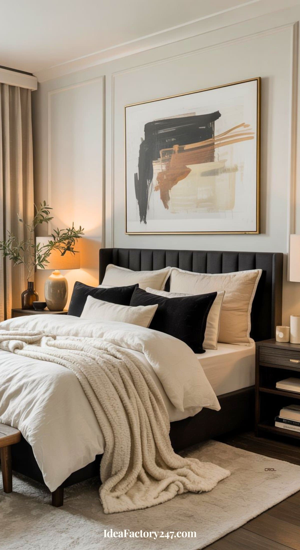A modern bedroom with a dark upholstered bed, white and beige bedding, black accent pillows, a cozy knit throw, a nightstand with a lamp, and an abstract painting above the bed. Natural light filters through beige curtains.