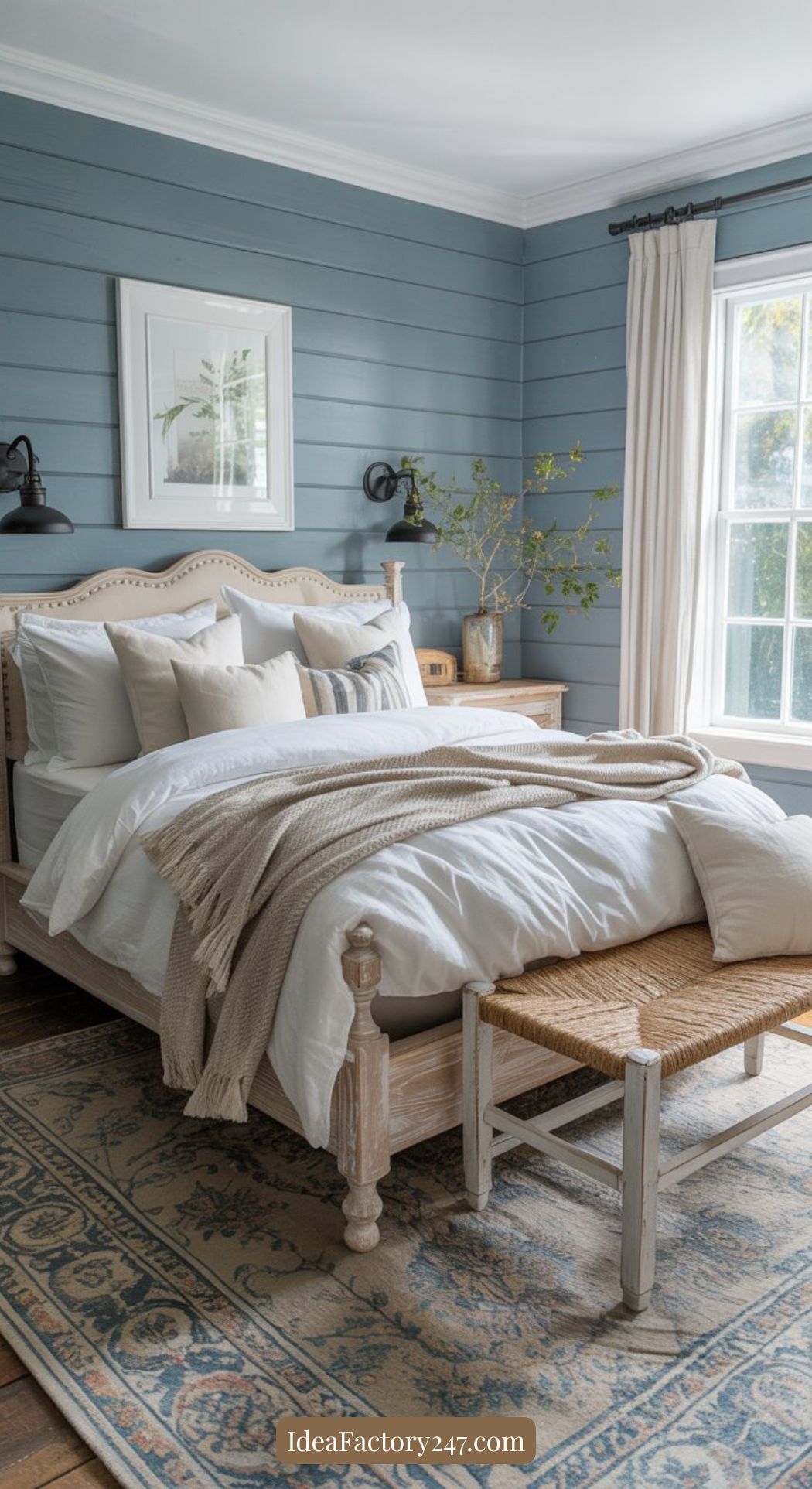 A cozy bedroom with a light wood bed, white bedding, beige throw, woven bench, blue paneled walls, large window with white curtains, wooden nightstand, plants, and a patterned rug on a hardwood floor.