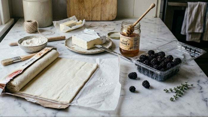 A marble countertop displays puff pastry dough, a bowl of flour, a stick of butter, cream cheese, a jar of honey with a dipper, fresh blackberries, sprigs of thyme, and a Honey & Pistachio Goat Cheese Log, ready for baking.