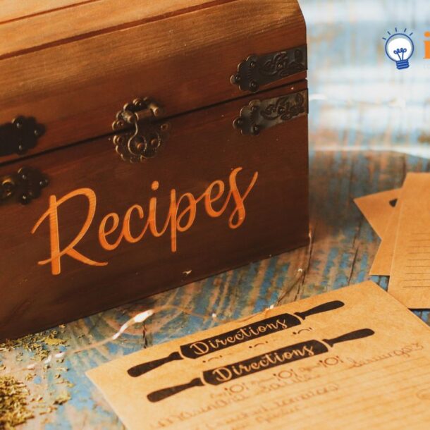 A wooden recipe box labeled Recipes sits next to handwritten recipes, spices, and measuring spoons on a rustic wooden table. The idea factory logo is in the top right corner.
