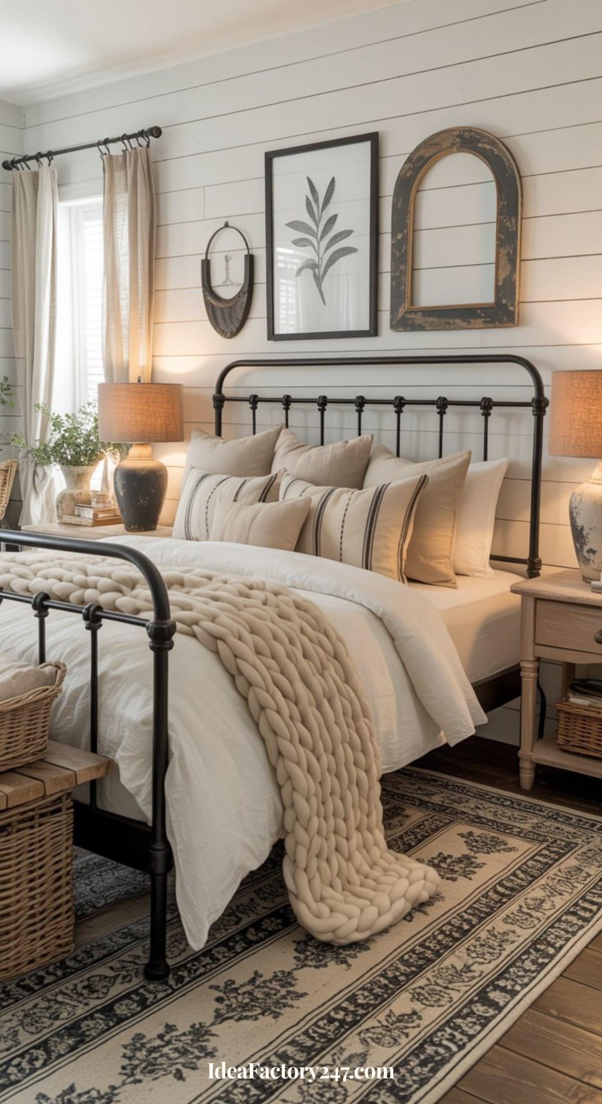 A cozy bedroom with a black metal bed frame, white bedding, beige throw pillows, a chunky knit blanket, wooden nightstands with lamps, baskets, and neutral decor on white shiplap walls, creating a rustic, inviting atmosphere.