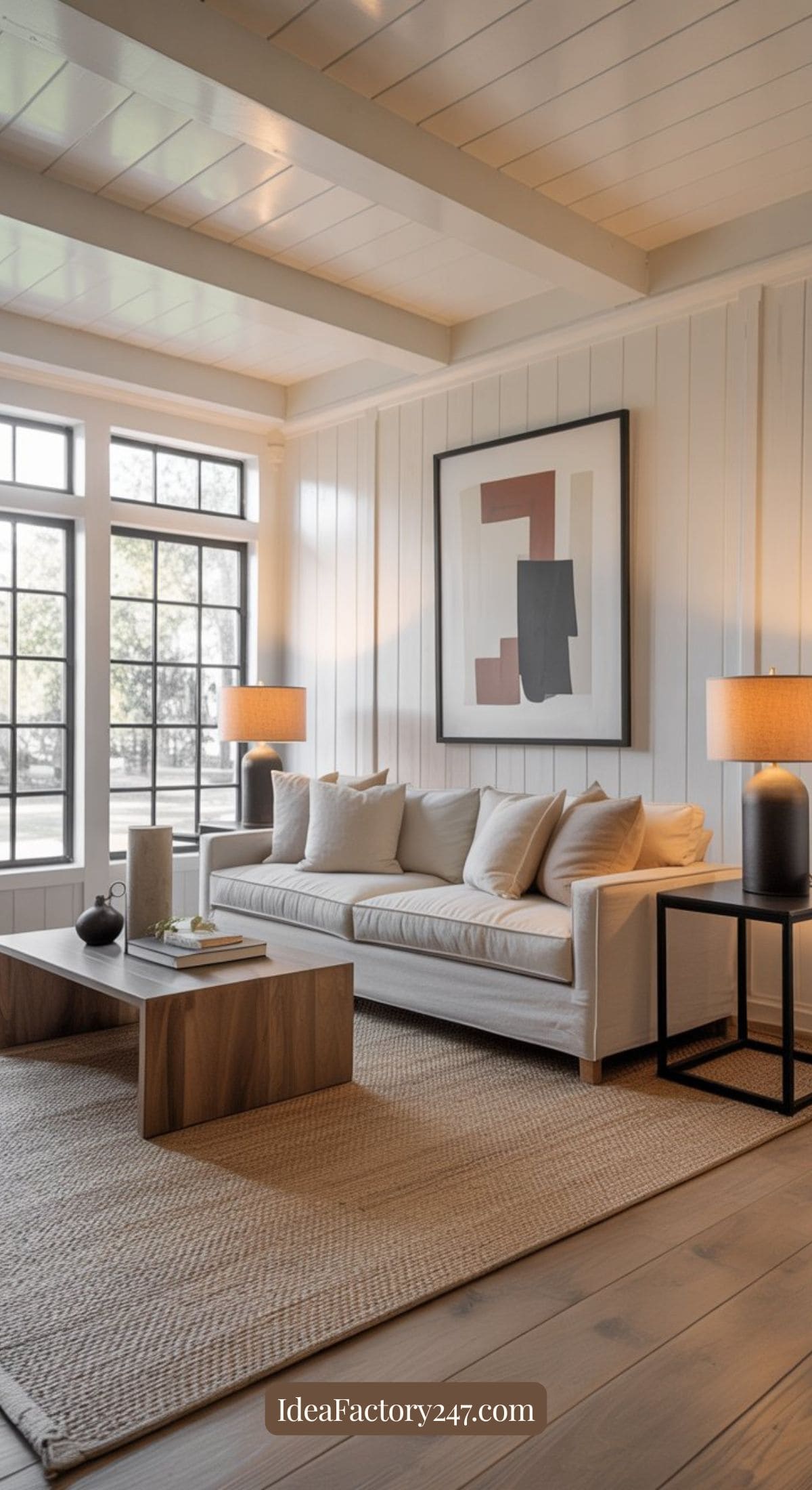 A modern living room with a beige sofa, neutral cushions, two wooden side tables, abstract wall art, table lamps, and large windows letting in natural light. A textured rug covers the wooden floor.