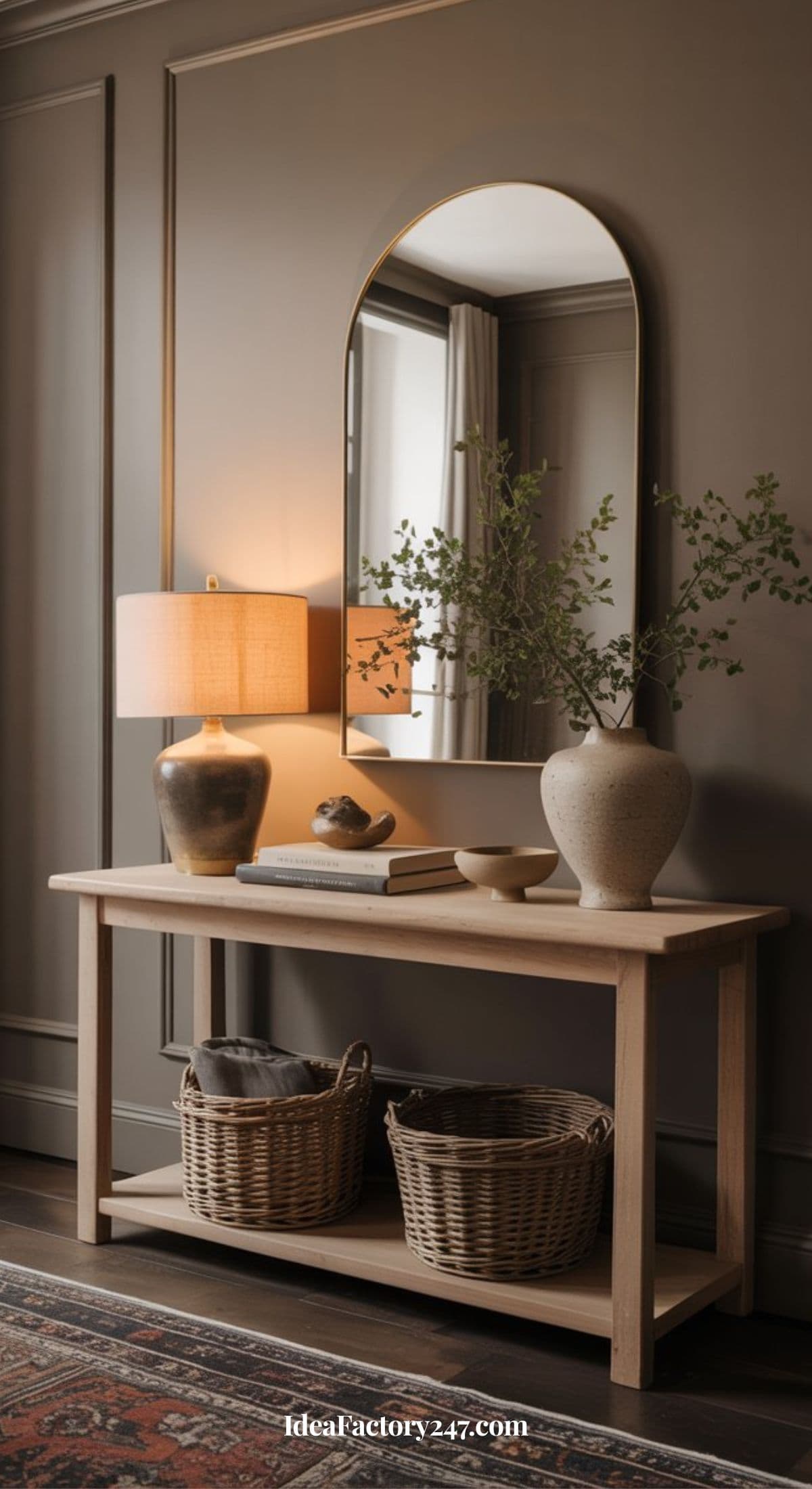 A wooden console table with two wicker baskets underneath, decorated with a lamp, books, a ceramic vase with greenery, and a sculptural object, stands against a gray paneled wall with a large arched mirror above.