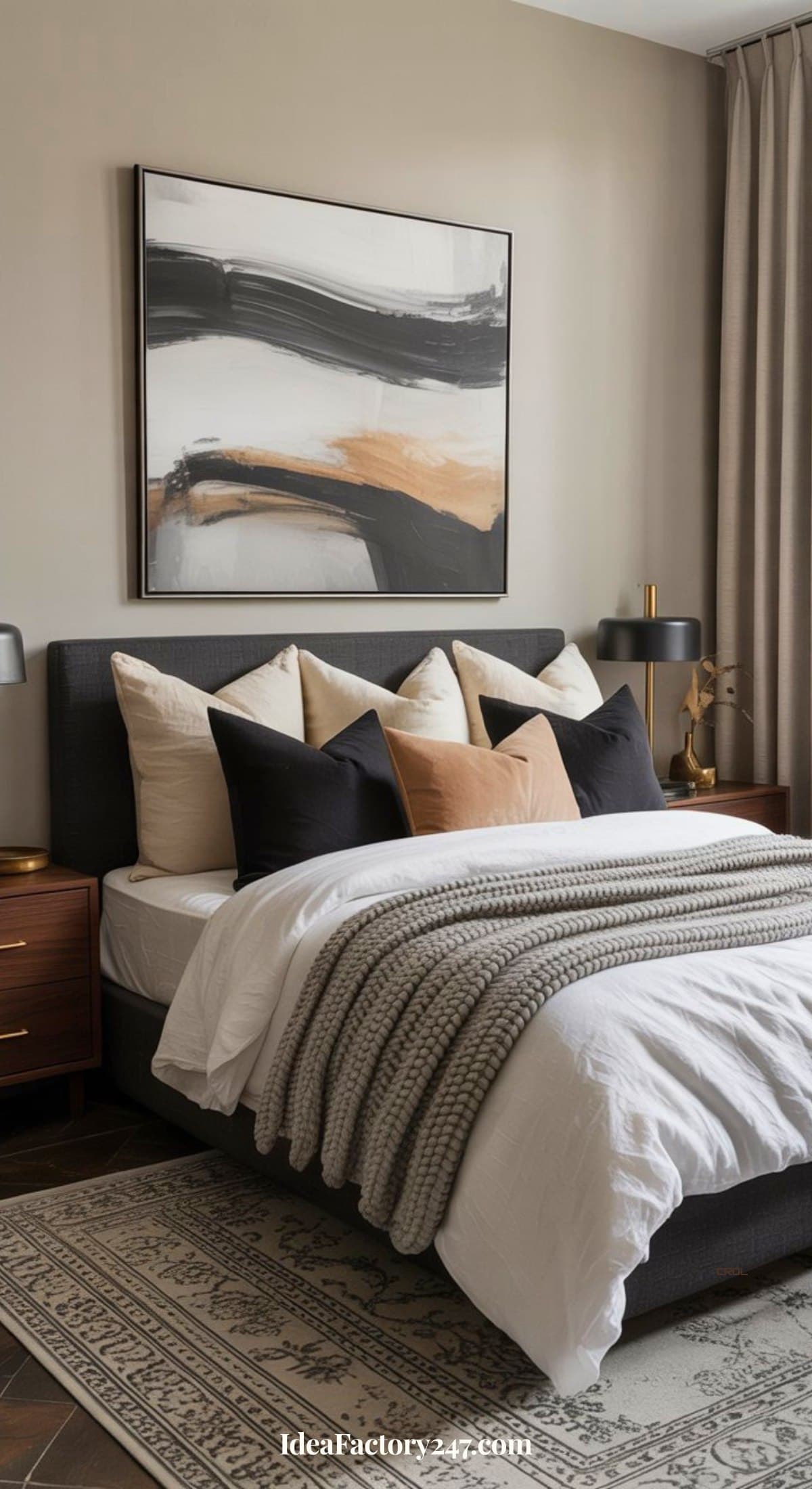 Modern bedroom with a dark upholstered bed, neutral bedding, knit blanket, and decorative pillows. Abstract art hangs above the bed; two wooden nightstands with black lamps sit on either side. A patterned rug covers the floor.