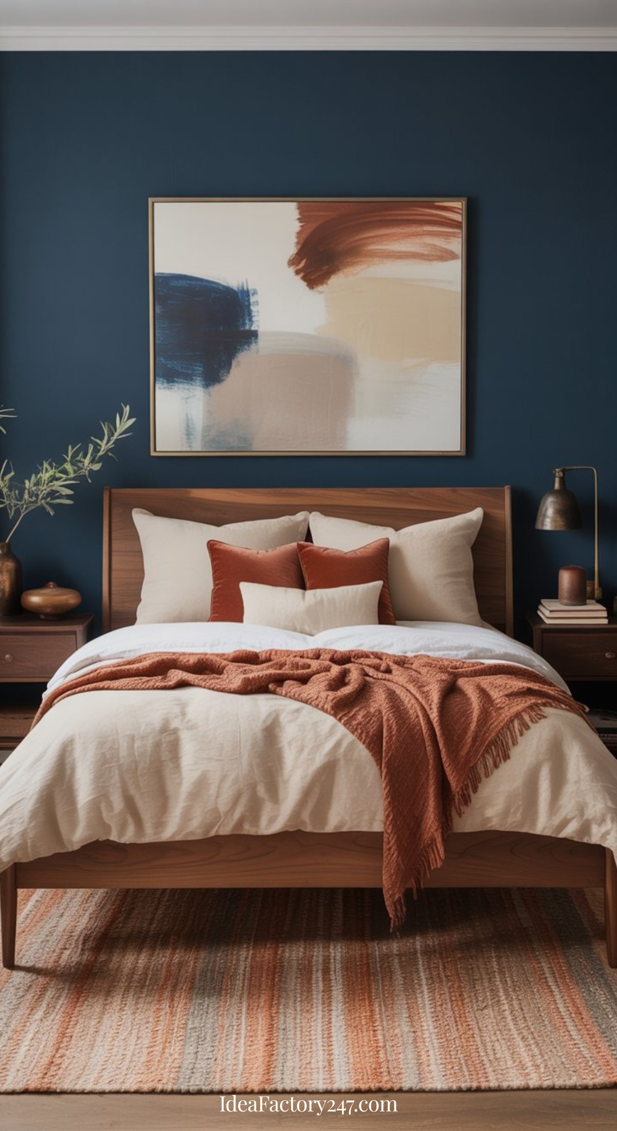 A cozy blue and terracotta bedroom features a wooden bed with cream and rust-colored bedding, matching pillows, and a rust throw. A modern abstract painting hangs on a dark blue wall, with nightstands and lamps on either side.