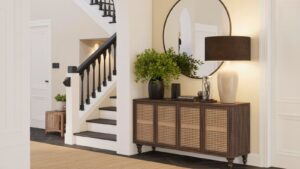 A modern entryway features a wooden console table with wicker panels, a large round mirror, a black table lamp, a green potted plant, and decor. A staircase with black railings is in the background.