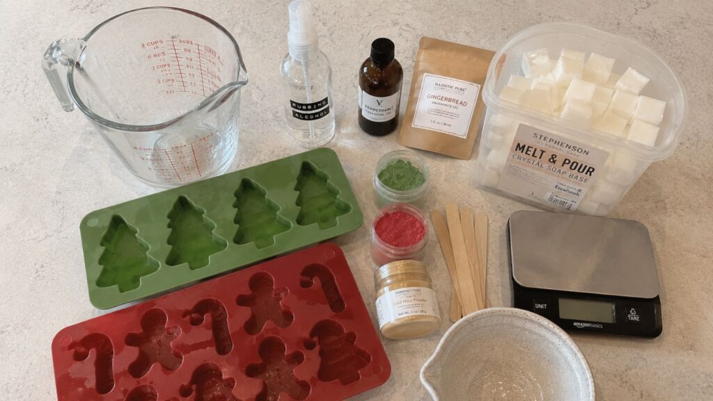 Soap-making supplies on a countertop, including a measuring cup, molds (tree and gingerbread shapes), fragrance oil, soap base cubes, color powders, digital scale, small bowl, spray bottle, wooden sticks, and a small jar.