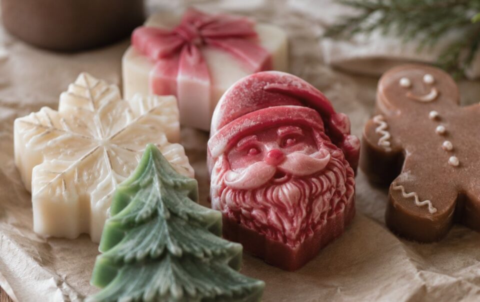 Assorted holiday-themed soaps shaped like a Christmas tree, Santa Claus, a snowflake, a present with a bow, and a gingerbread figure, displayed on crumpled paper.