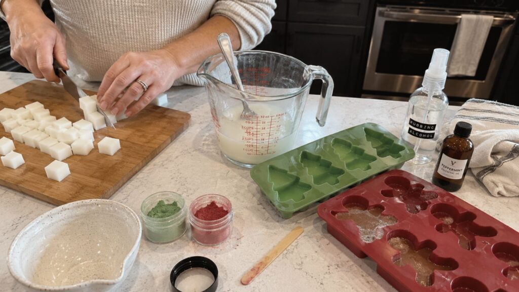 Person chopping soap base cubes on a cutting board, with soap-making supplies, molds, color powders, fragrance, and measuring cup on a kitchen counter.