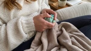 A person in a cream sweater sits on a couch holding a fidget toy and a beige blanket, with a mug and notebook on a nearby table.