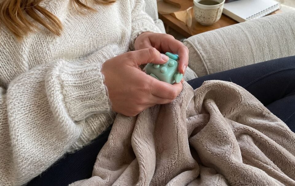 A person in a cream sweater sits on a couch holding a fidget toy and a beige blanket, with a mug and notebook on a nearby table.
