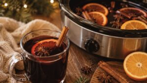 A glass mug of mulled wine garnished with an orange slice, cinnamon stick, and star anise sits next to a slow cooker filled with more, surrounded by oranges, cinnamon, and festive decor.