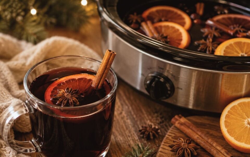 A glass mug of mulled wine garnished with an orange slice, cinnamon stick, and star anise sits next to a slow cooker filled with more, surrounded by oranges, cinnamon, and festive decor.