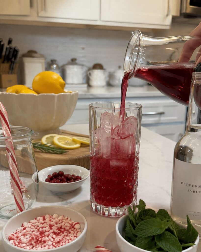 A hand pours red liquid into a tall glass with ice. Around are lemons, peppermint syrup, crushed peppermints, cranberries, a candy cane, mint leaves, and lemon slices on a white kitchen counter.