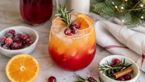 A festive Sunrise Mocktail Recipe with orange and red layers, garnished with cranberries, an orange slice, and rosemary. Surrounded by fresh cranberries, orange, and a cinnamon stick on a marble surface.