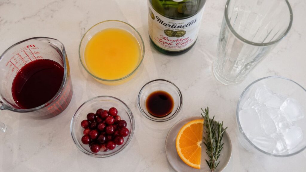 Ingredients for a Sunrise Mocktail Recipe on a counter: measuring cup with red liquid, bowl of orange juice, sparkling cider bottle, glass of ice, small bowl of cranberries, small bowl of dark liquid, orange slice, and rosemary sprig.