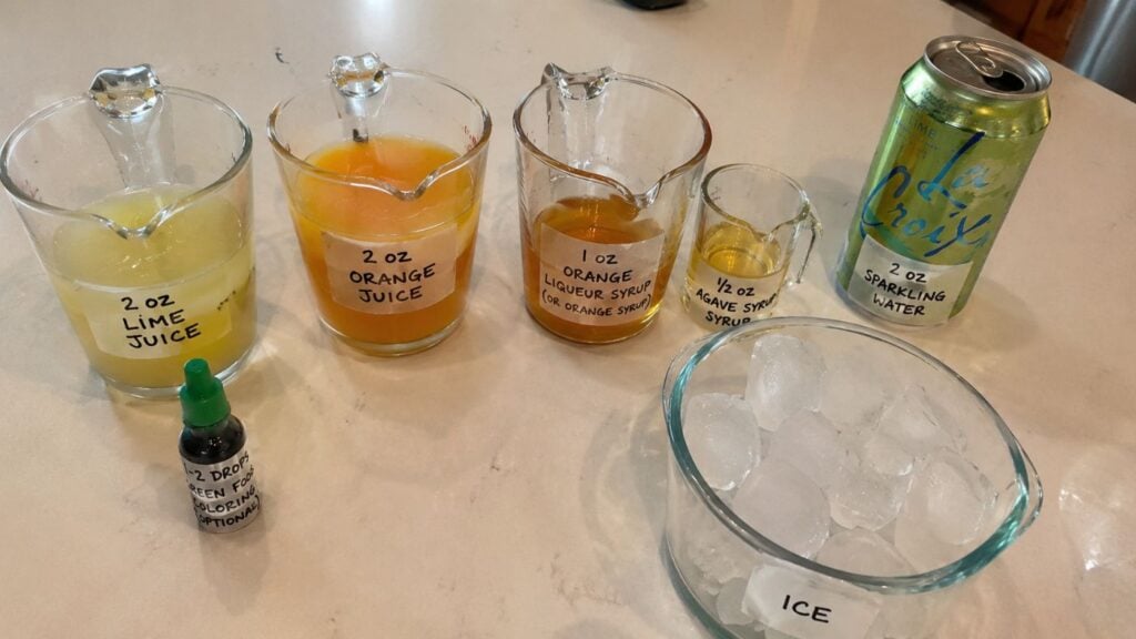 Ingredients for a drink: lime juice, orange juice, orange liqueur syrup, agave syrup, green stevia, sparkling water can, and a bowl of ice, all labeled and arranged on a countertop.