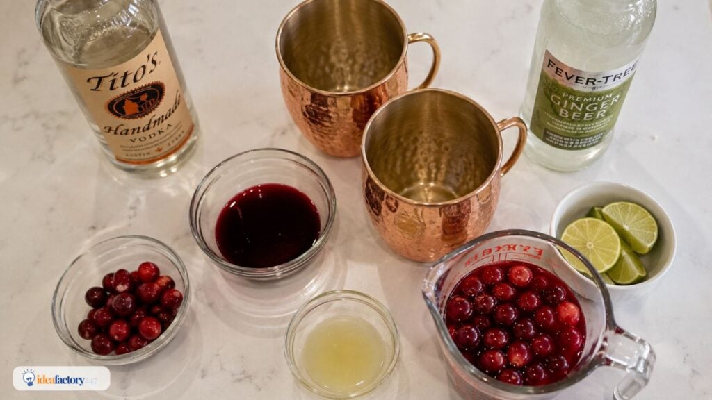 Ingredients for a cranberry mule cocktail on a counter: vodka, ginger beer, copper mugs, cranberry juice, cranberries, lime wedges, and lime juice in glass containers.