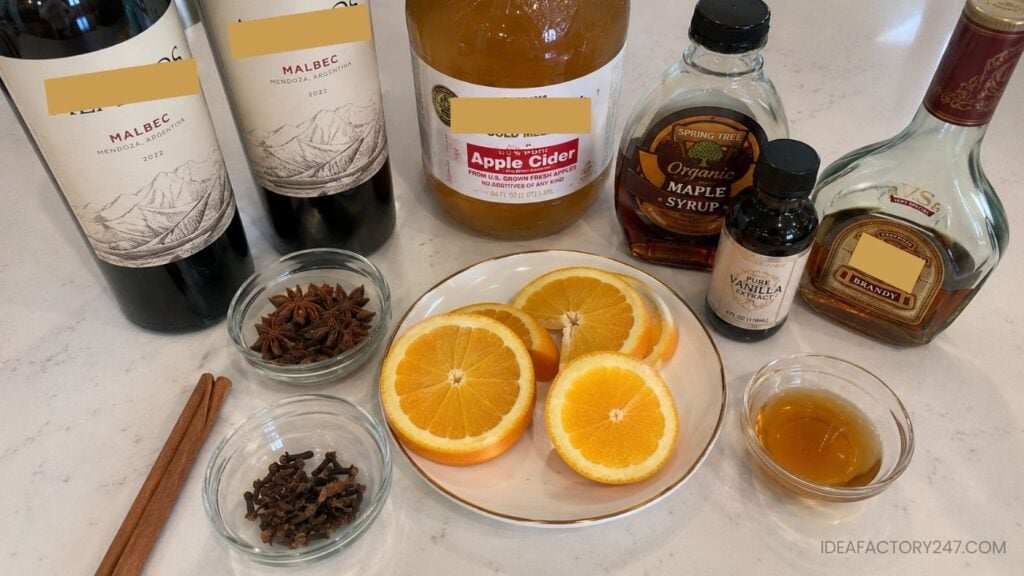Various ingredients on a counter: two bottles of Malbec wine, apple cider, maple syrup, vanilla extract, orange slices, honey, cinnamon stick, star anise, and cloves in small bowls.