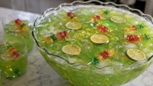 A punch bowl filled with bright green drink, ice cubes, lime slices, and skewered gummy bears. Two glasses with the same drink and garnishes are beside the bowl on a marble surface.