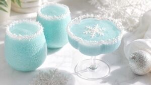 Three light blue frozen drinks in festive glasses with snowflake decorations and white crystal rims, set on a white surface with silver ornaments and snowflakes nearby.