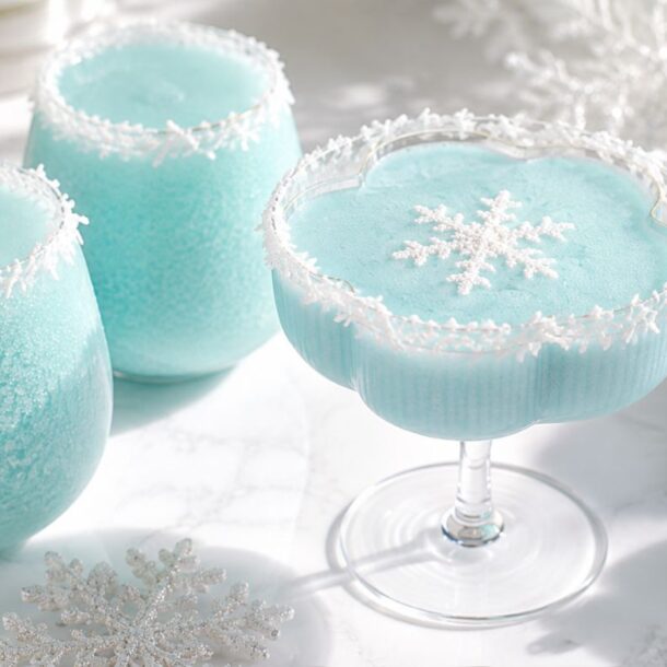 Three light blue frozen drinks in festive glasses with snowflake decorations and white crystal rims, set on a white surface with silver ornaments and snowflakes nearby.