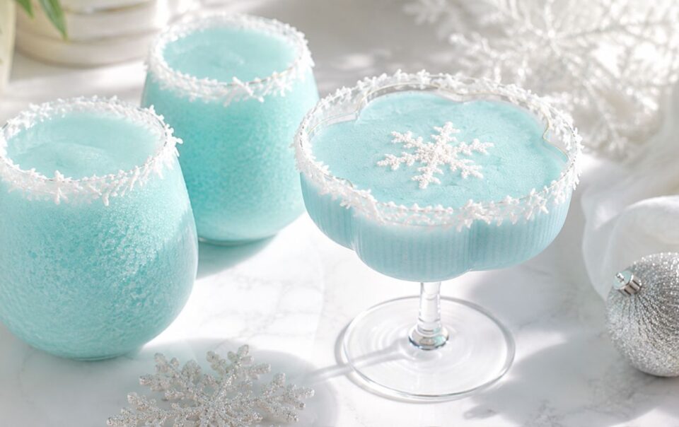Three light blue frozen drinks in festive glasses with snowflake decorations and white crystal rims, set on a white surface with silver ornaments and snowflakes nearby.