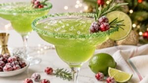 A green margarita in a glass with a green sugar rim, garnished with a lime slice, cranberries, and rosemary. Christmas decorations and sugared cranberries are in the background.