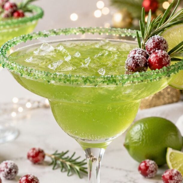 A green margarita in a glass with a green sugar rim, garnished with a lime slice, cranberries, and rosemary. Christmas decorations and sugared cranberries are in the background.