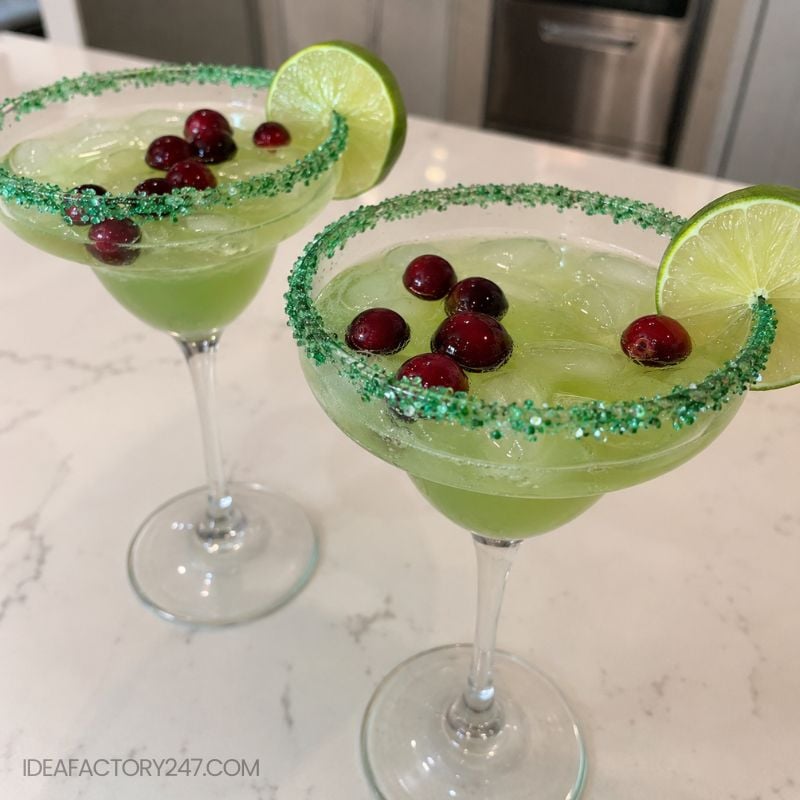 Two margarita glasses with green drinks, green sugared rims, ice, fresh cranberries, and lime slices garnish, set on a white counter.