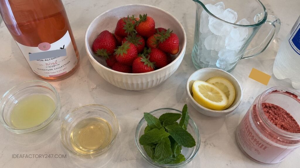 A bowl of strawberries, a pitcher of ice, lemon slices, mint, lemon juice, a bottle of rosé, and a container of pink drink powder are arranged on a white counter—perfect ingredients for crafting a refreshing Pink Mocktail Recipe.