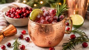A copper mug filled with a cranberry cocktail, ice, sugared cranberries, a lime wedge, and a rosemary sprig. Cranberries, rosemary, and a bowl of cranberries are on the marble surface nearby.