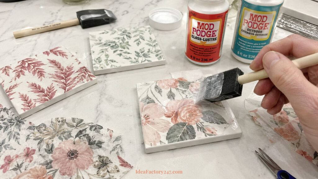 A hand applies Mod Podge to a floral-patterned tile with a foam brush. Other tiles, Mod Podge bottles, scissors, and floral napkins are on a white surface.