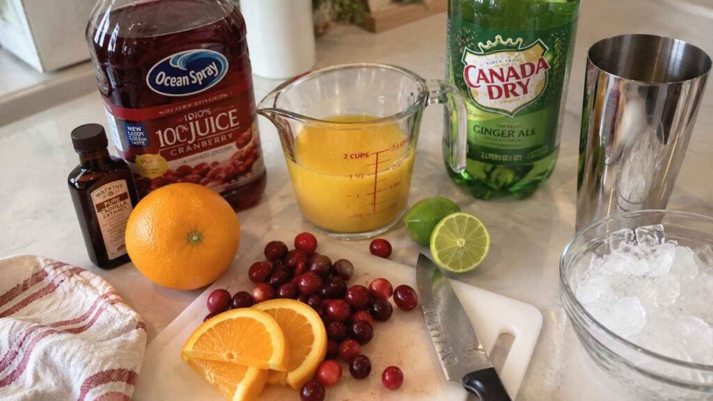 Ingredients for a festive Cranberry Vanilla Crush Holiday Mocktail, including cranberry juice, orange juice, ginger ale, vanilla, fresh orange and lime, cranberries, a knife, ice, and a cocktail shaker, are arranged on a kitchen counter.