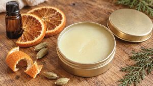 A tin of balm with its lid off sits on a wooden surface next to dried orange slices, orange peel, a small brown bottle, green cardamom pods, and evergreen sprigs—showcasing the inviting ingredients perfect for creating your own DIY Solid Perfume.