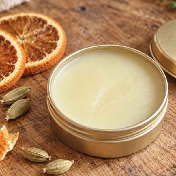 A tin of balm with its lid off sits on a wooden surface next to dried orange slices, orange peel, a small brown bottle, green cardamom pods, and evergreen sprigs—showcasing the inviting ingredients perfect for creating your own DIY Solid Perfume.