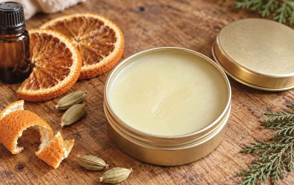 A tin of balm with its lid off sits on a wooden surface next to dried orange slices, orange peel, a small brown bottle, green cardamom pods, and evergreen sprigs—showcasing the inviting ingredients perfect for creating your own DIY Solid Perfume.