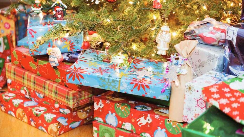 Wrapped Christmas presents in colorful paper are stacked under a decorated and lit Christmas tree, with ornaments, lights, and garlands visible on the branches above the gifts.
