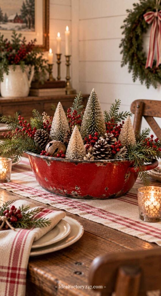 A rustic holiday table features a red bowl with mini snow-covered trees, pinecones, and berries. Candles, greenery, and festive linens enhance the cozy, Christmas-themed decor.