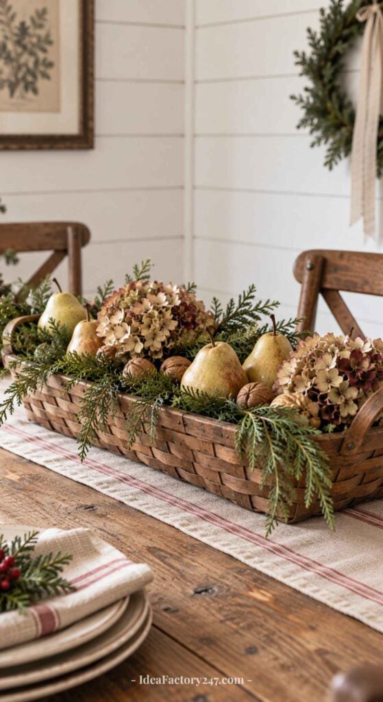A woven basket centerpiece on a wooden table, filled with pears, walnuts, dried hydrangea flowers, and evergreen branches. The table has a striped runner, and a wreath hangs on the white wall in the background.