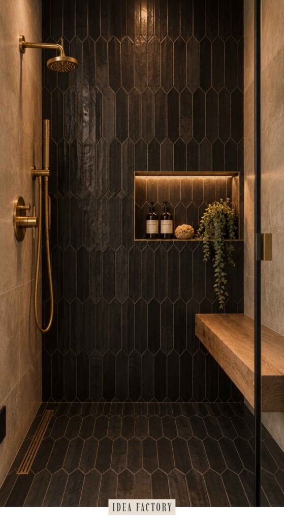 A walk-in shower with dark hexagonal wall tiles, brass fixtures, a floating wooden bench, and a recessed shelf holding two bottles and a trailing plant.