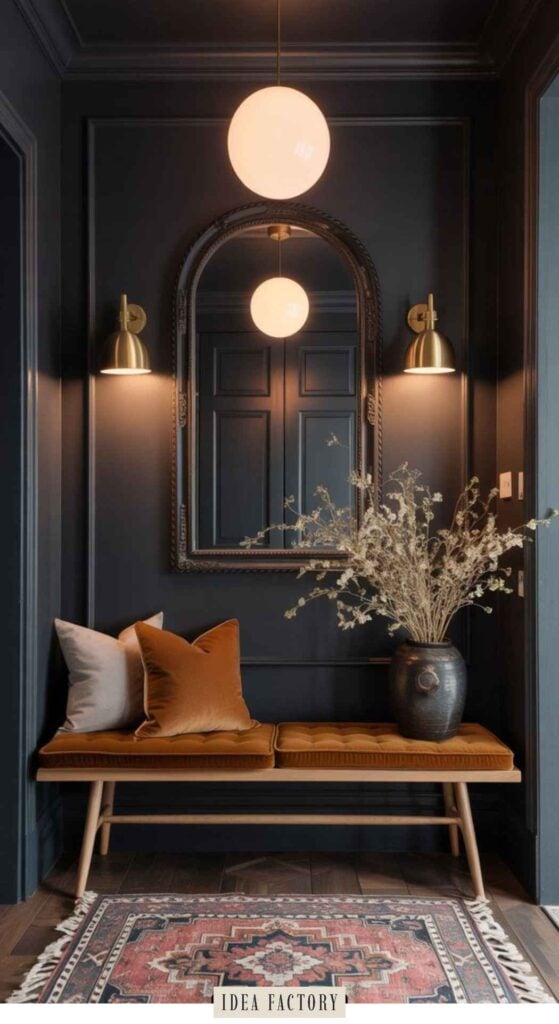 A bench with cushions and a vase of dried flowers sits beneath an arched mirror and wall sconces in a dark-painted entryway.