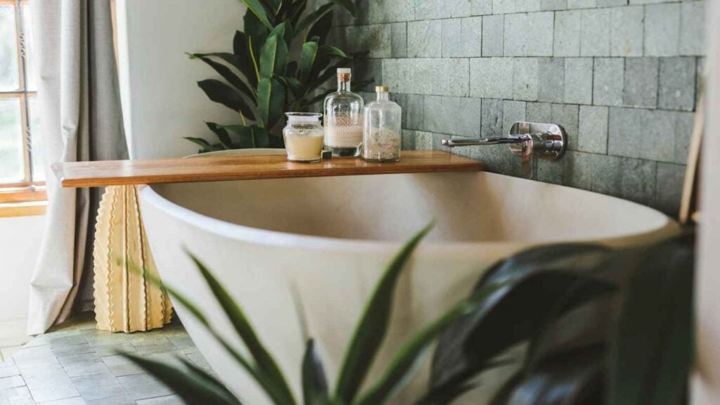 A freestanding bathtub with a wooden tray holding three glass jars, set against a green tiled wall decorated with bathroom plants.