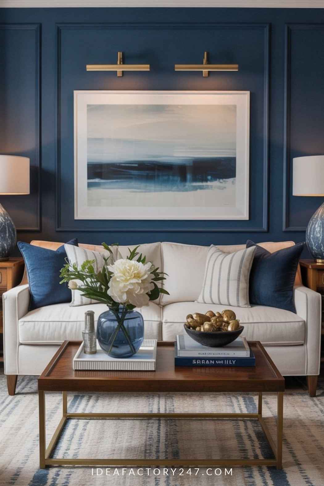 A white sofa with blue and striped pillows sits against a dark blue paneled wall beneath a large landscape painting and two gold wall lights, with a coffee table holding a vase of flowers and a bowl of gold spheres in the foreground.